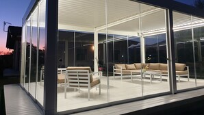 A modern glass-enclosed outdoor living area with a sofa and chairs, a wooden deck, and a view of the surrounding landscape. - Il Rifugio Del Tempo (Roccasecca)