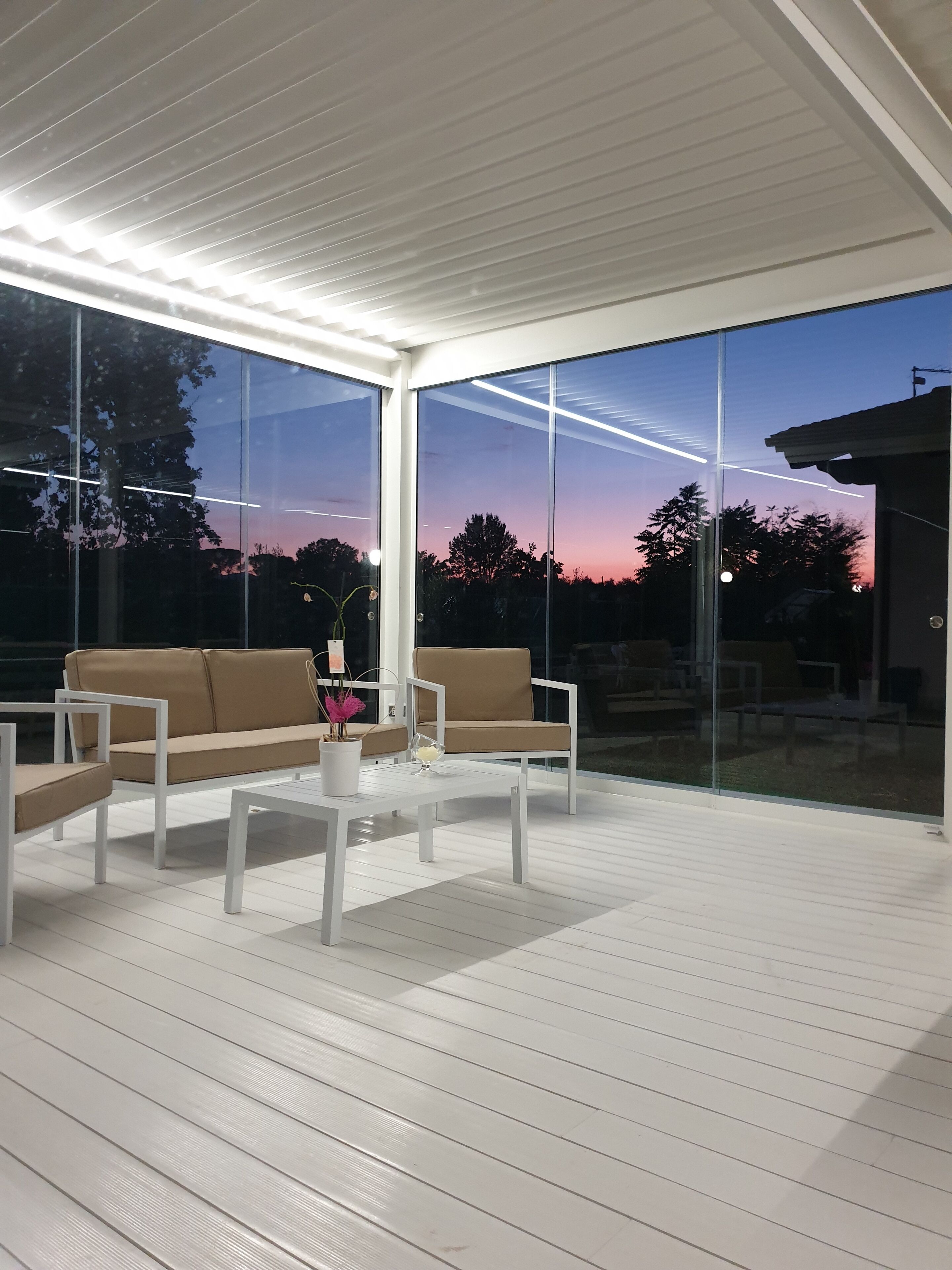 Ein moderner Außenbereich zum Sitzen mit weißer Möbel und großen Glasfenstern, der Blick auf den Himmel und Bäume bietet.