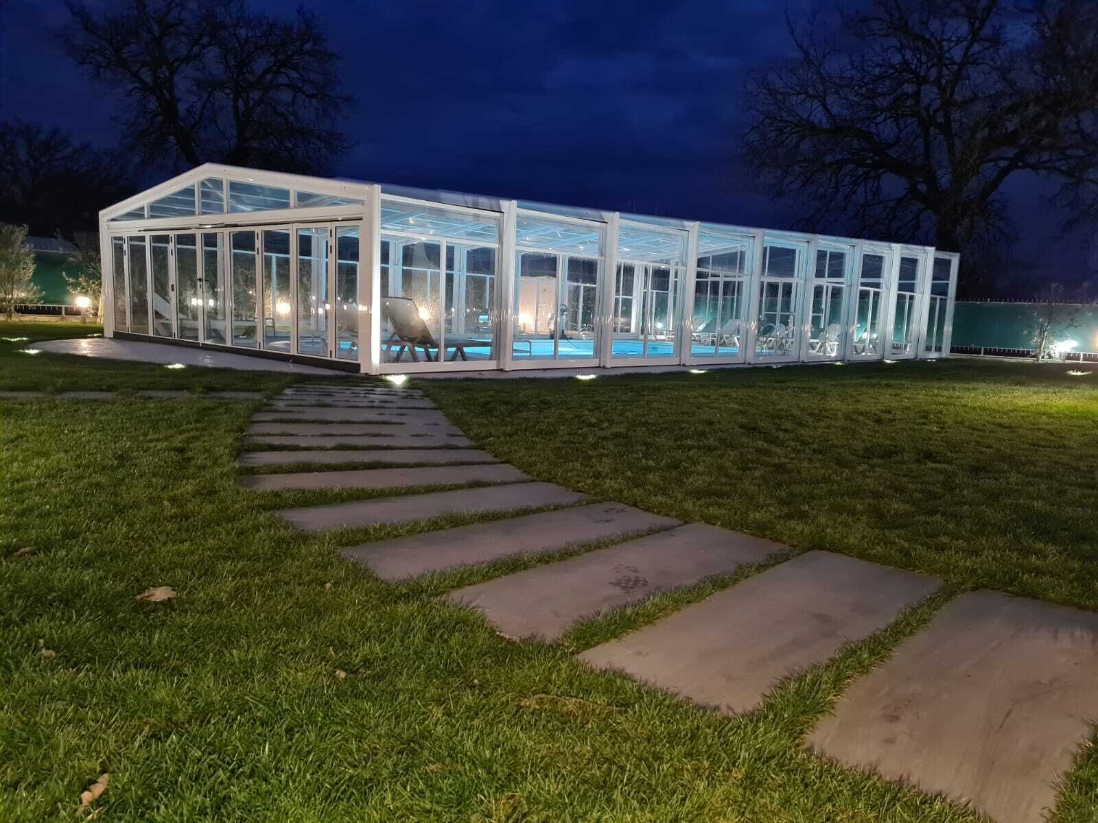 A glass-enclosed structure with a pool, surrounded by a stone pathway and grass.