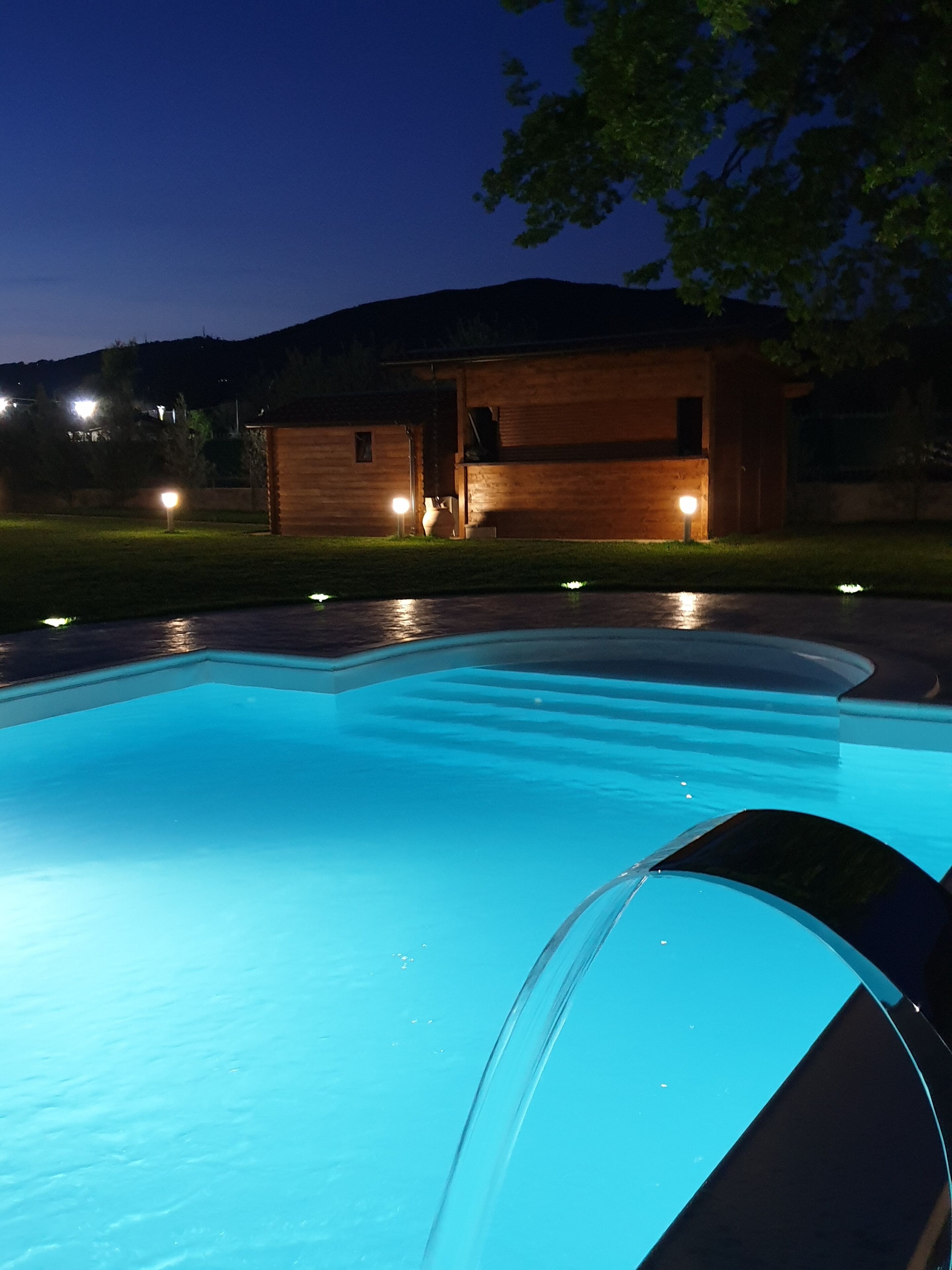 Una piscina de vora corbada, envoltada d'un pont de fusta i un edifici petit amb llums.
