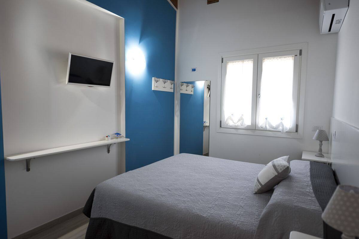 A bedroom with a bed, a TV mounted on the wall, a window with curtains, and a bedside table with a lamp.