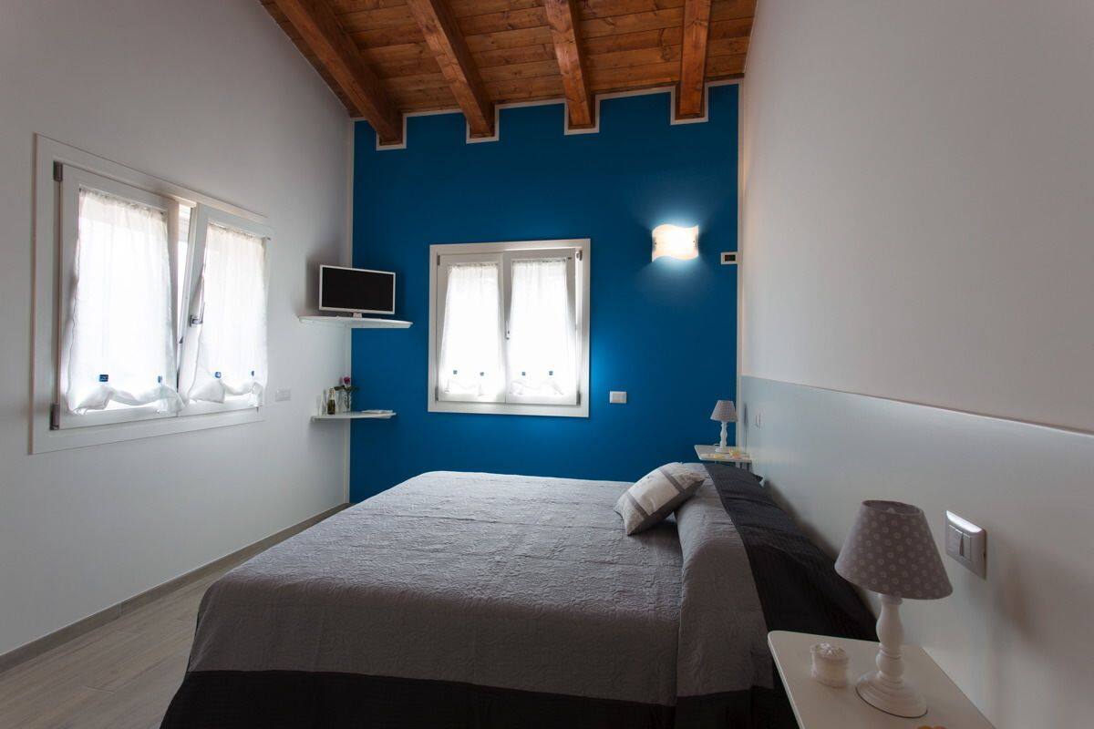 A bedroom with a bed, two windows, a TV, and a nightstand with a lamp.