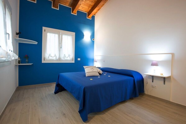 Une pièce avec un mur bleu, un lit recouvert d’une couvre-lit bleue, une fenêtre avec des rideaux blancs, une table de chevet avec une lampe et une étagère avec un vase.