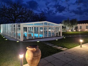 A glass-enclosed pool area with a clear view of the pool, surrounded by a well-lit patio and a large urn in the foreground. - Il Rifugio Del Tempo (Roccasecca)