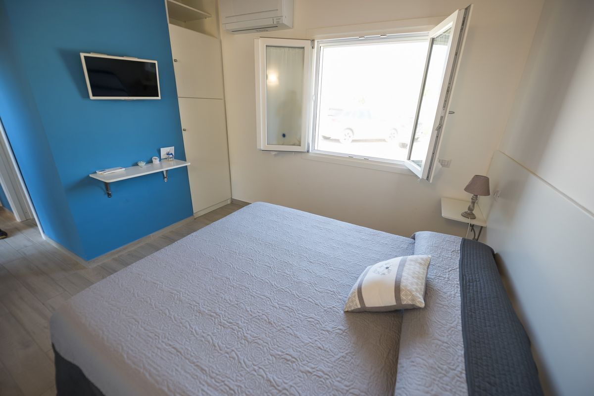 A bedroom with a bed, a bedside table with a lamp, a window, and a TV on the wall.