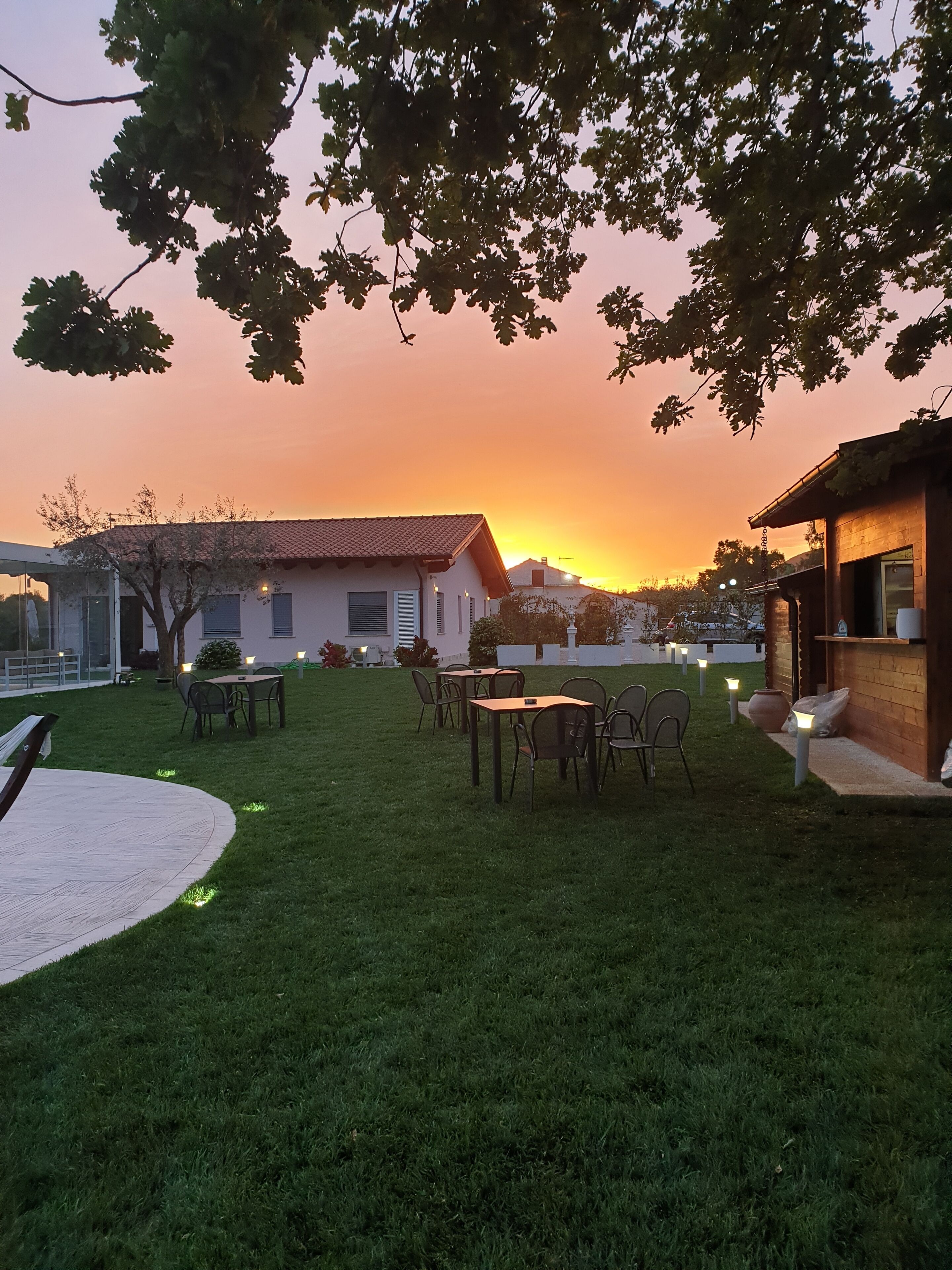A grassy outdoor area with tables and chairs, a building with a tiled roof, and a sunset in the background.