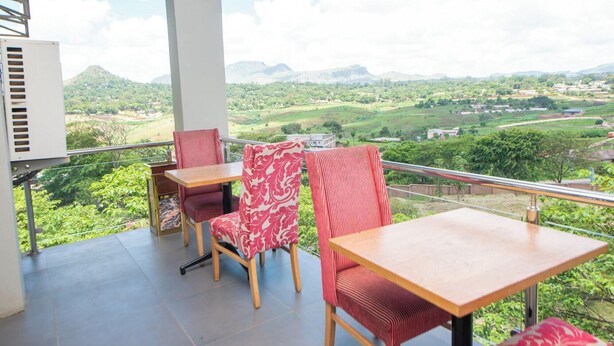 A balcony with two wooden tables and two red upholstered chairs, overlooking a scenic landscape with hills and buildings.