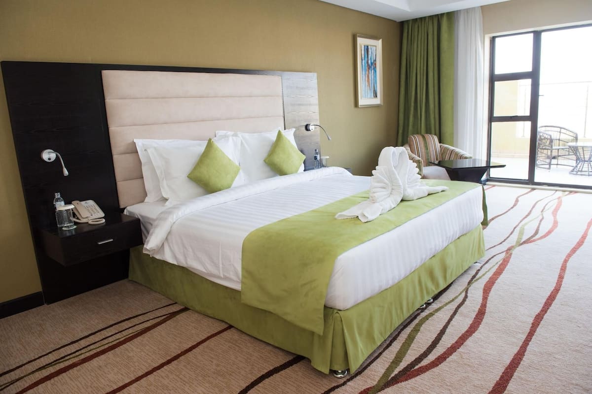 A hotel room with a large bed, a bedside phone, a green throw blanket, and a view of the outdoors through a window.