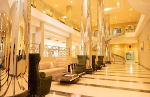 A spacious lobby with a high ceiling, a central seating area with sofas and armchairs, a staircase leading to an upper level, and a glass railing overlooking the lobby. - Crossroads Hotel Blantyre (Blantyre)
