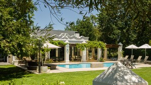 A pool area with lounge chairs and umbrellas, surrounded by greenery and a building with columns.