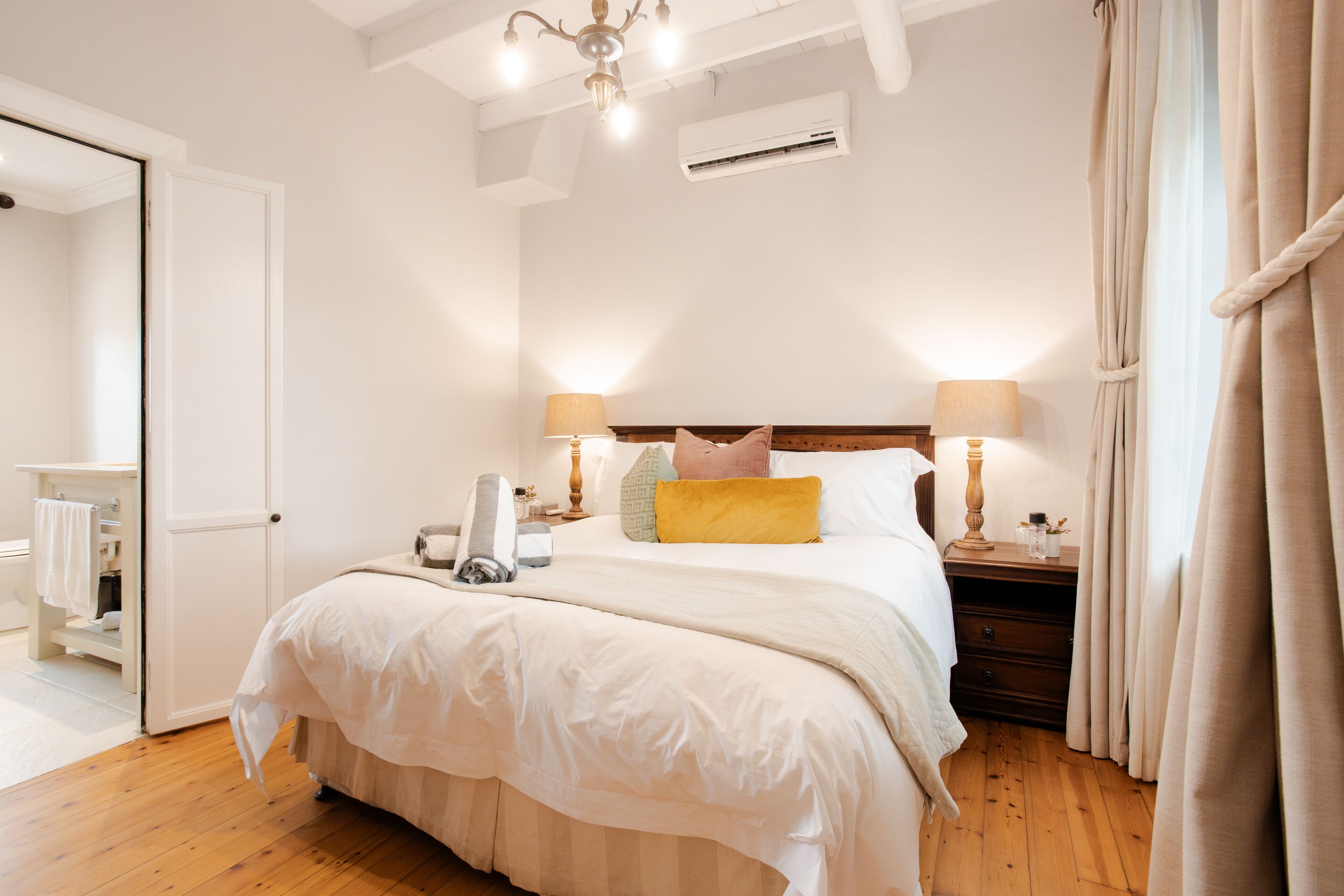 Un dormitorio con suelo de madera, una cama con ropa blanca, dos mesitas de noche con lámparas y un ventilador de techo.