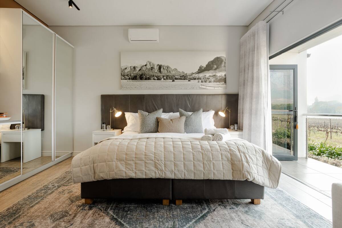 A modern bedroom with a large bed, bedside tables, a mounted artwork, and a view through a glass door.