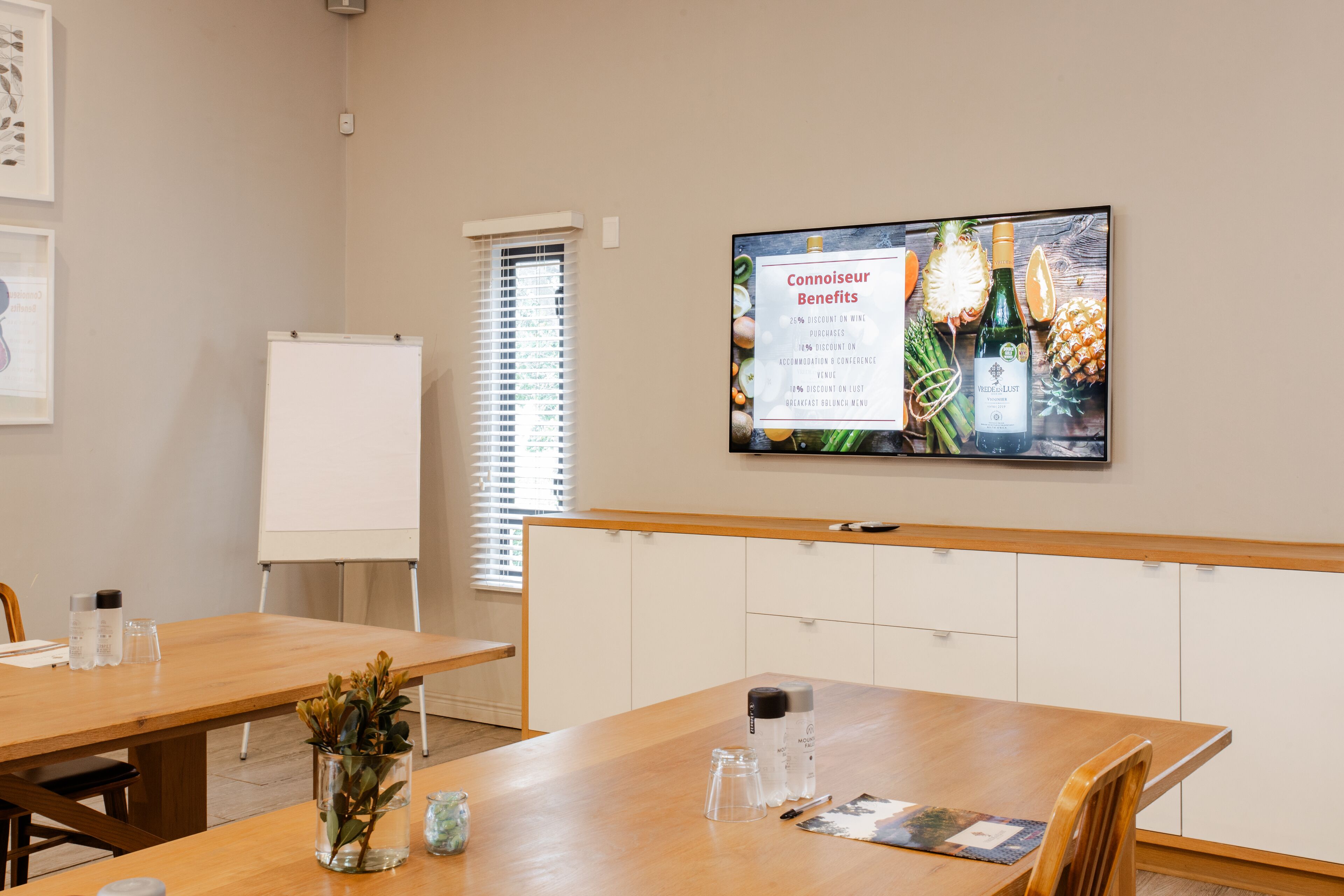 A conference room with a whiteboard, a large screen displaying benefits, and a wooden table with various items.