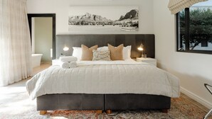 A bedroom with a large bed, a headboard, a bedside table with a lamp, a window with a view, and a bathroom with a bathtub and toilet.