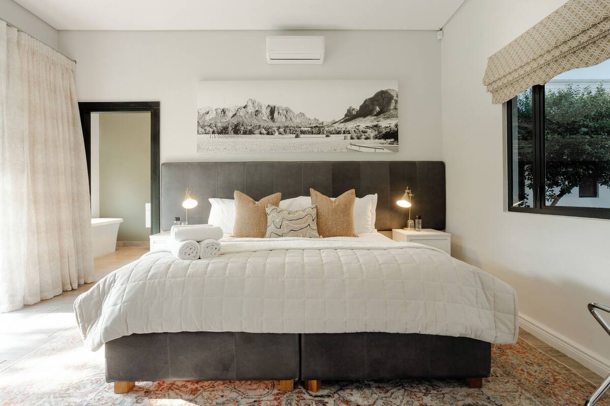 A bedroom with a large bed, a headboard, a bedside table with a lamp, a window with a view, and a bathroom with a bathtub and toilet.