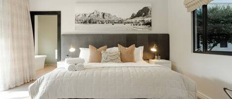 Une chambre avec un grand lit, une tête de lit, une table de chevet avec une lampe, une fenêtre donnant sur un beau paysage, et une salle de bain avec une baignoire et des toilettes.