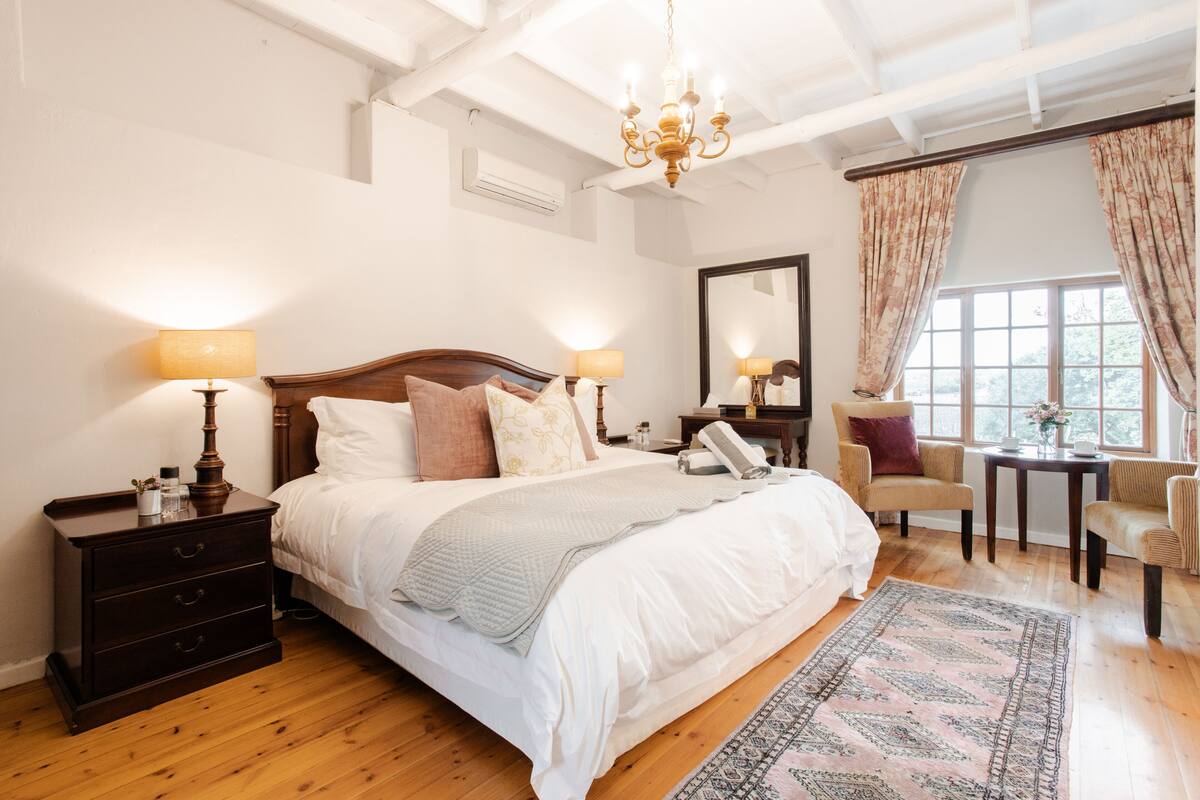 A bedroom with a large bed, bedside lamps, a nightstand, a chair, a small table, and a window with curtains.