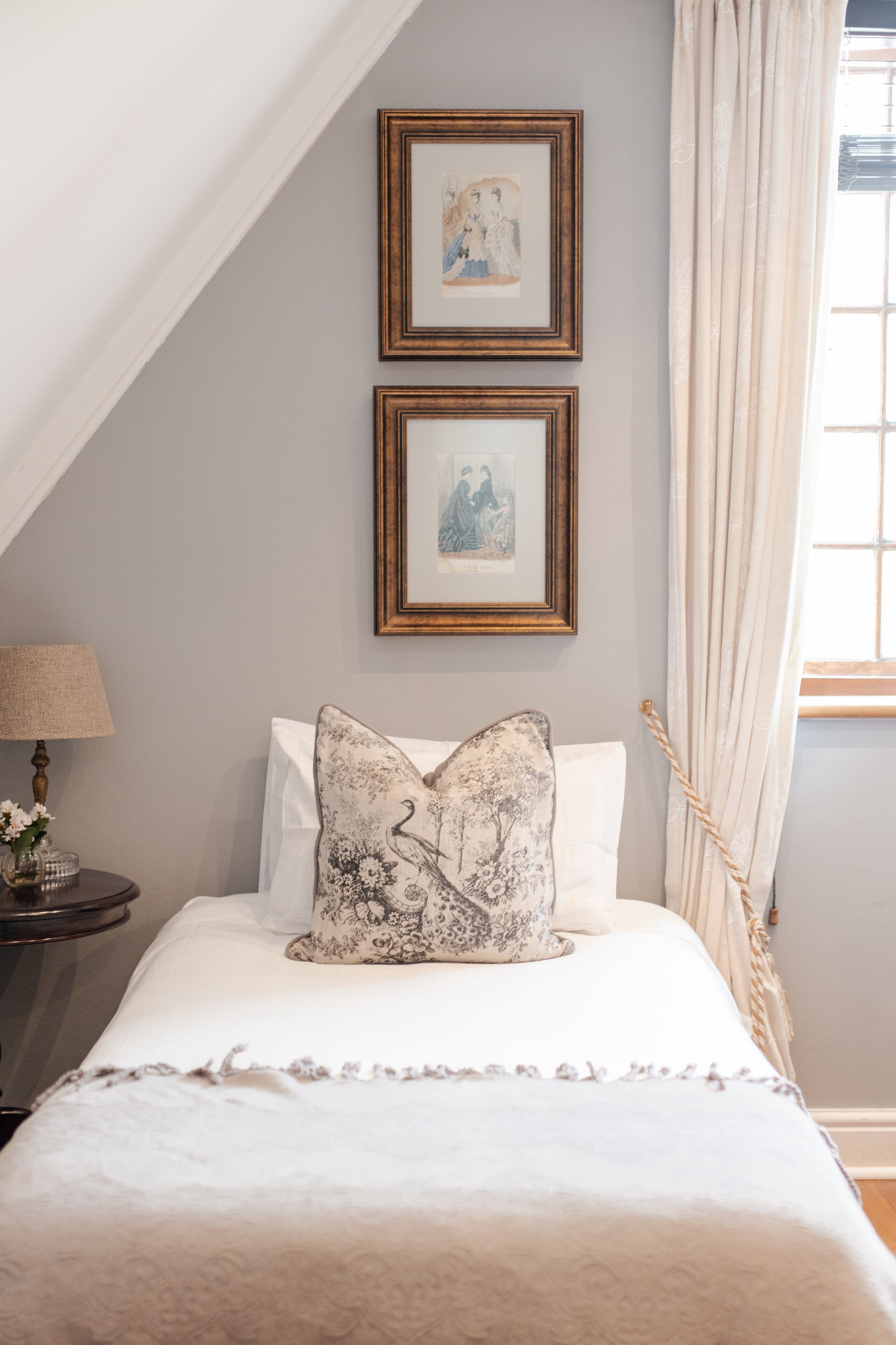 A neatly made bed with white linens and decorative pillows, framed artwork on the wall, and a bedside table with a lamp.