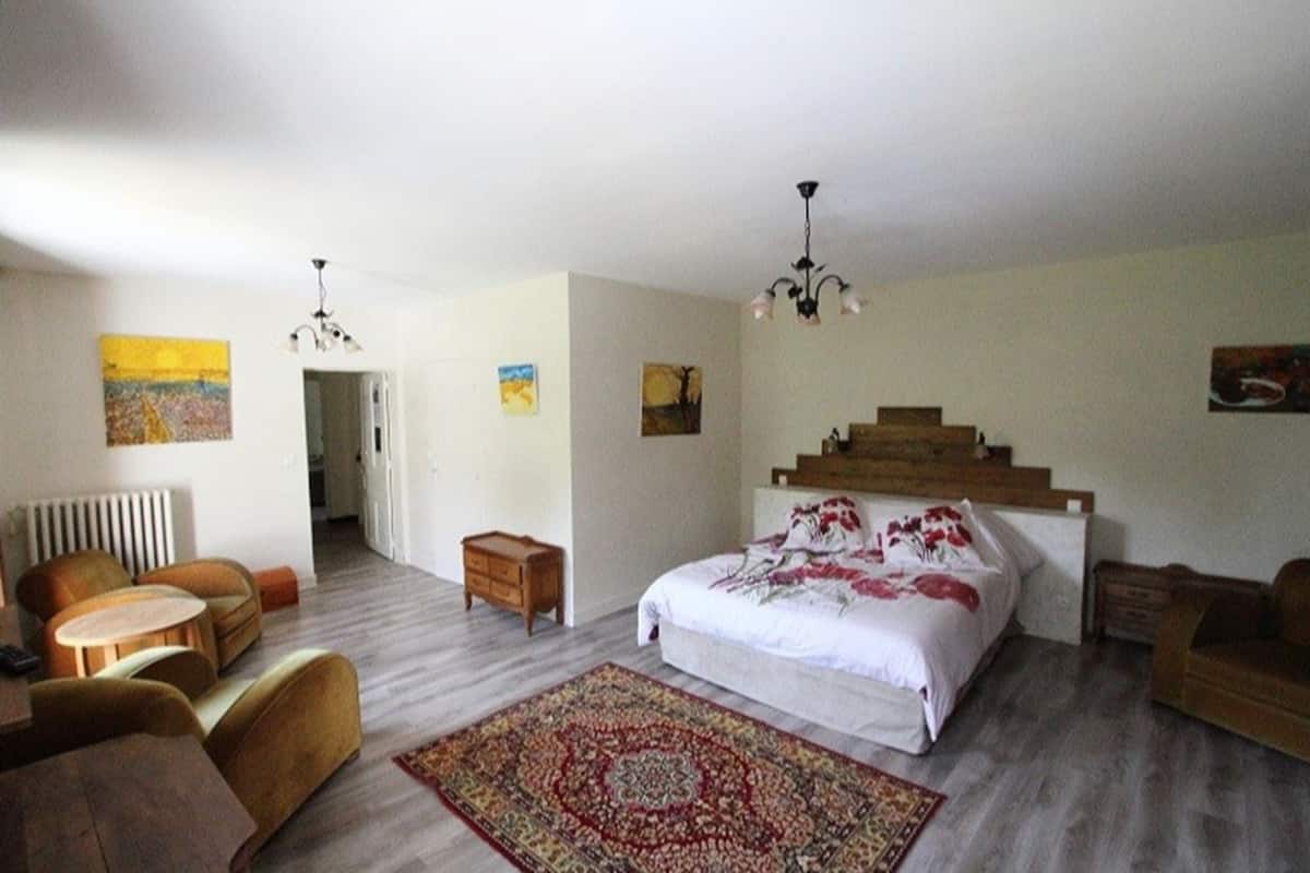 Un dormitorio amplio con una cama grande, cabecera de madera, un rincón de descanso con una silla y una mesa, y una alfombra estampada.