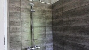 Área de banho com divisória de vidro, paredes revestidas de azulejos e chuveiro de metal.