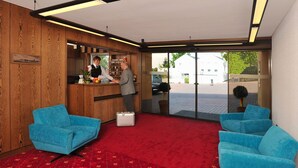 Zona de recepción de hotel con un mostrador de madera, dos sillones azules, una alfombra roja y un amplio ventanal hacia el exterior.
