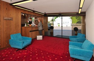 Uma área de recepção de hotel com um balcão de madeira, duas poltronas azuis, um tapete vermelho e uma grande porta de vidro que dá para o exterior.