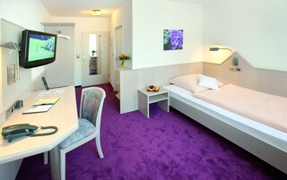 Quarto de hotel com uma cama, uma escrivaninha com cadeira, uma televisão, uma mesinha com um vaso de flores e um quadro na parede.