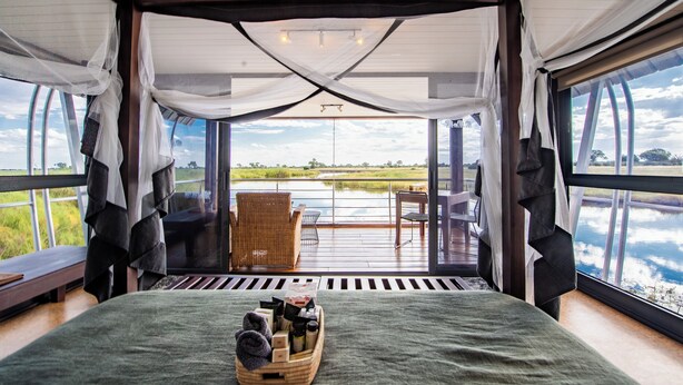 A room with a bed, a wicker chair, a table, and a view of a body of water.