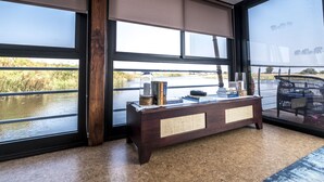 Ein Zimmer mit großen Fenstern und Blick auf ein Gewässer, ein Holzkonsolentisch mit Büchern und Dekoration sowie ein Korbstuhl.