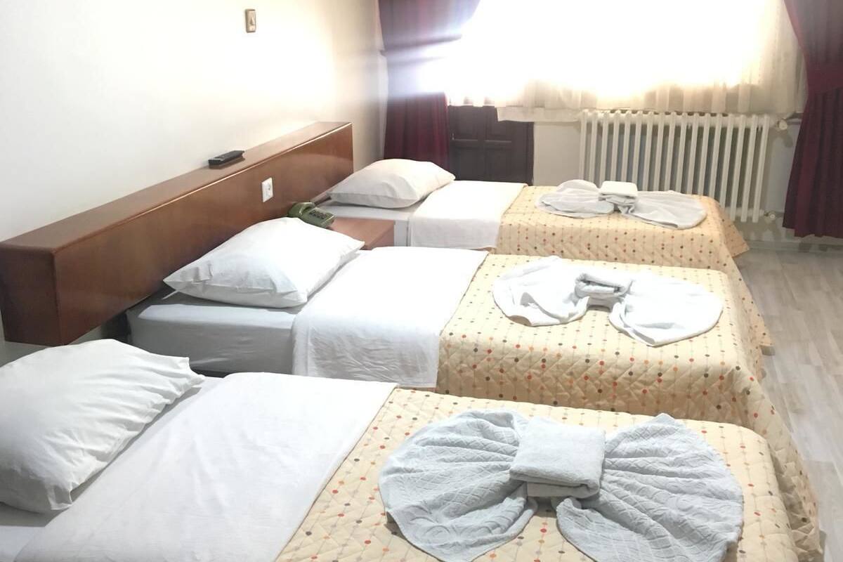 Ein Hotelzimmer mit drei Einzelbetten, jedes mit einem weißen Kopfkissen und einer Decke. Die Betten sind mit gemusterten Bettdecken ausgestattet. Das Bett links hat ein Holz-Kopfteil, und im Hintergrund ist ein Heizkörper zu sehen.