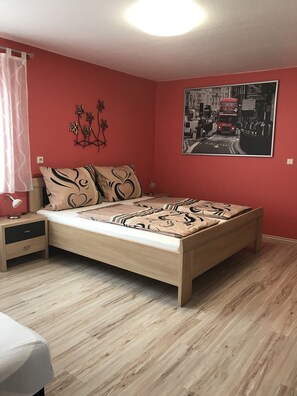 A bedroom with a wooden bed, a nightstand, a lamp, a red wall, and a black and white poster. - Gästehaus Durst (Rust)