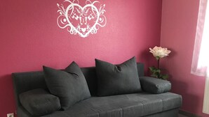 A grey sofa with multiple cushions against a pink wall, a white decorative heart, and a single white rose on the right. - Gästehaus Durst (Rust)