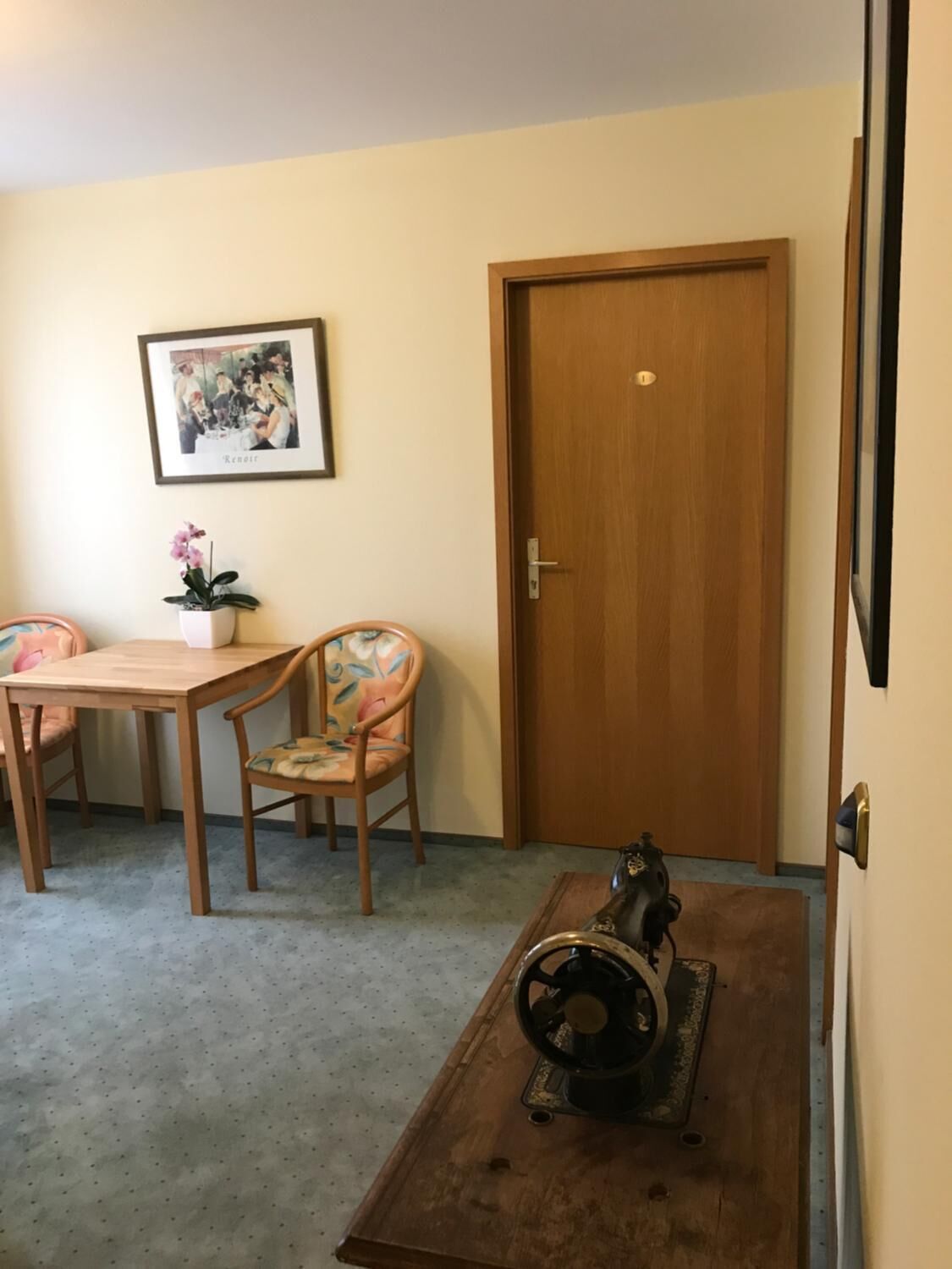 Ein Zimmer mit Holztür, einem gerahmten Bild an der Wand, einem Tisch mit Stühlen und einer Nähmaschine auf einem Holztisch.
