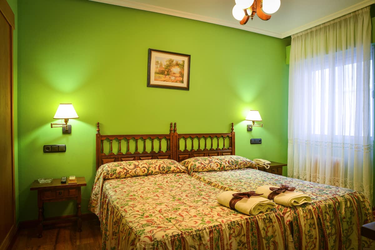 A room with two beds, green walls, a wooden headboard, two bedside tables with lamps, a framed picture on the wall, and a window with sheer curtains.
