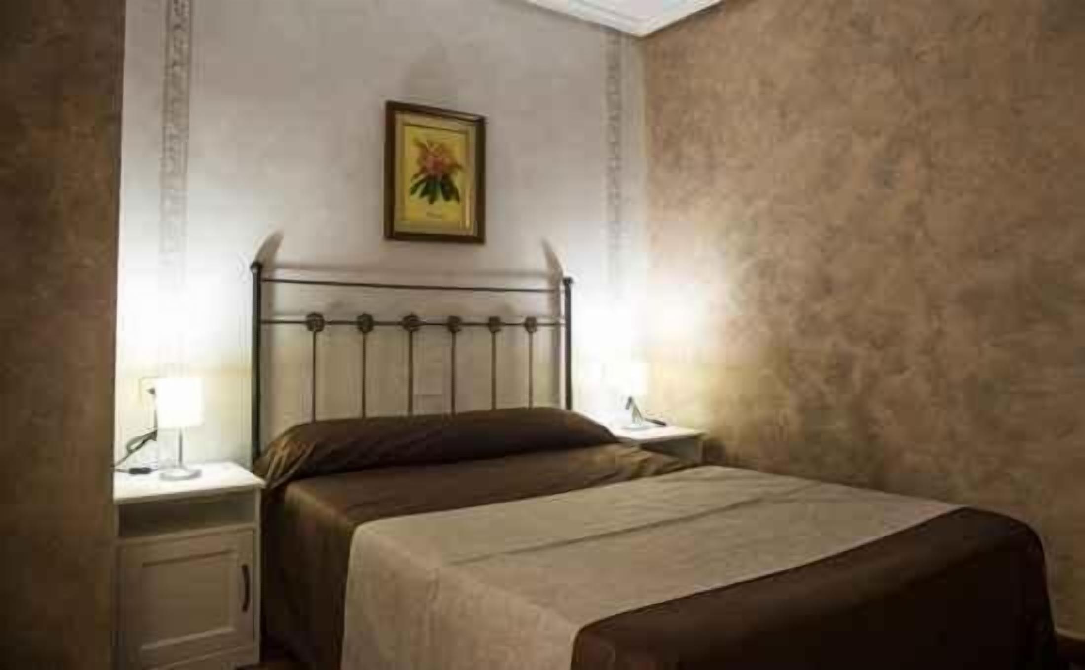 Une chambre à coucher avec un lit en métal, un lit simple recouvert d’une couverture marron, deux tables de chevet avec des lampes et un tableau encadré accroché au mur.