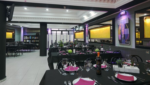 Une salle de banquet où les tables sont dressées pour le repas, avec des nappes noires, des assiettes blanches et des serviettes roses.
