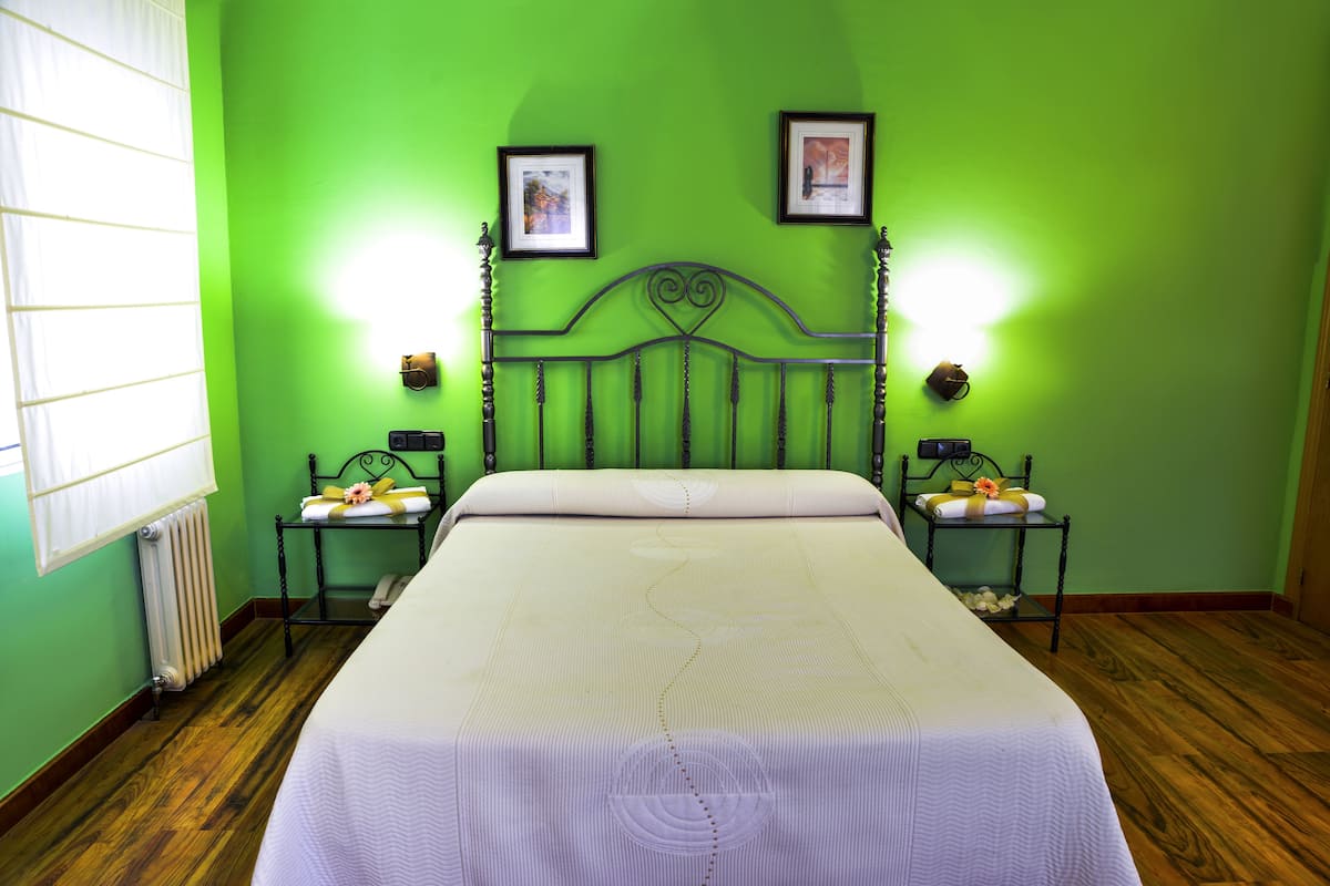 A room with a green wall, a wooden floor, a bed with a white bedspread, two bedside tables with lamps, and two framed pictures on the wall.