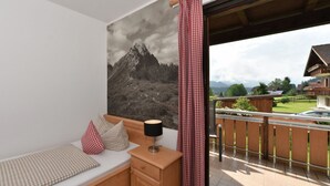 A balcony with a mountain view, a bed with checkered pillows, and a wooden nightstand with a lamp. - Landhaus Marga (Fischen im Allgaeu)