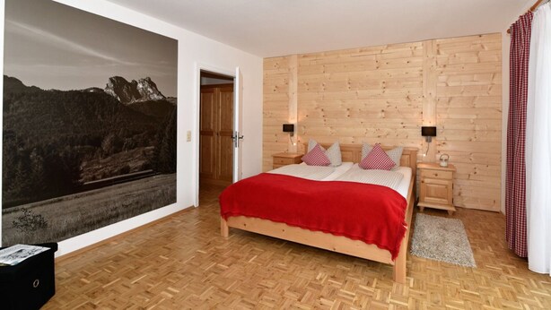 A bedroom with a wooden bed, a nightstand, a red and white checkered pillow, a wooden wall, and a window with a curtain. - Landhaus Marga (Fischen im Allgaeu)