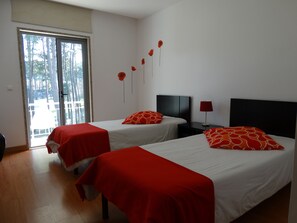 Ein Hotelzimmer mit zwei Betten, jeweils mit einer roten und weißen Bettdecke, einem Nachttisch mit einer Lampe und einer Schiebetür zu einem Außenbereich.