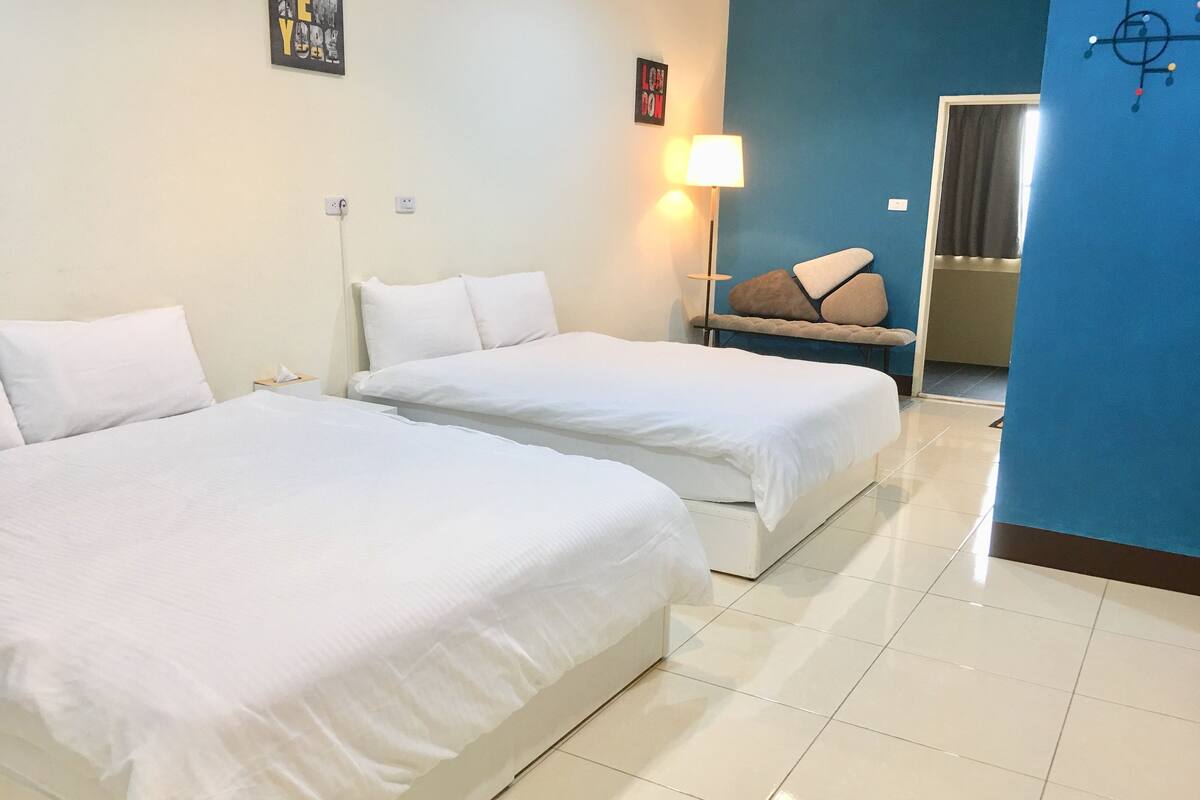 A hotel room with two beds, a nightstand, a lamp, and a blue accent wall.