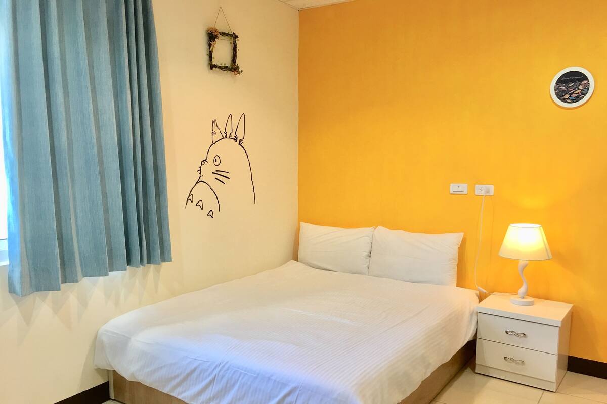 A bedroom with a bed, a nightstand, a lamp, a wall decoration, and a window with curtains.