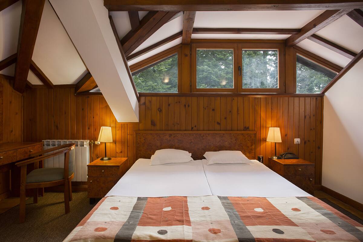 Een kamer met een houten plafond, twee bedden met witte lakens, een bureau en twee bedlampjes.