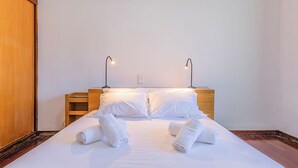 Ein ordentlich bezogenes Bett mit weißen Laken und zusammengerollten Handtüchern, zwei Nachttischlampen und ein hölzernes Kopfteil.