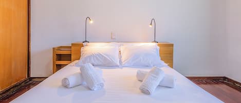 A neatly made bed with white linens and rolled towels, two bedside lamps, and a wooden headboard.
