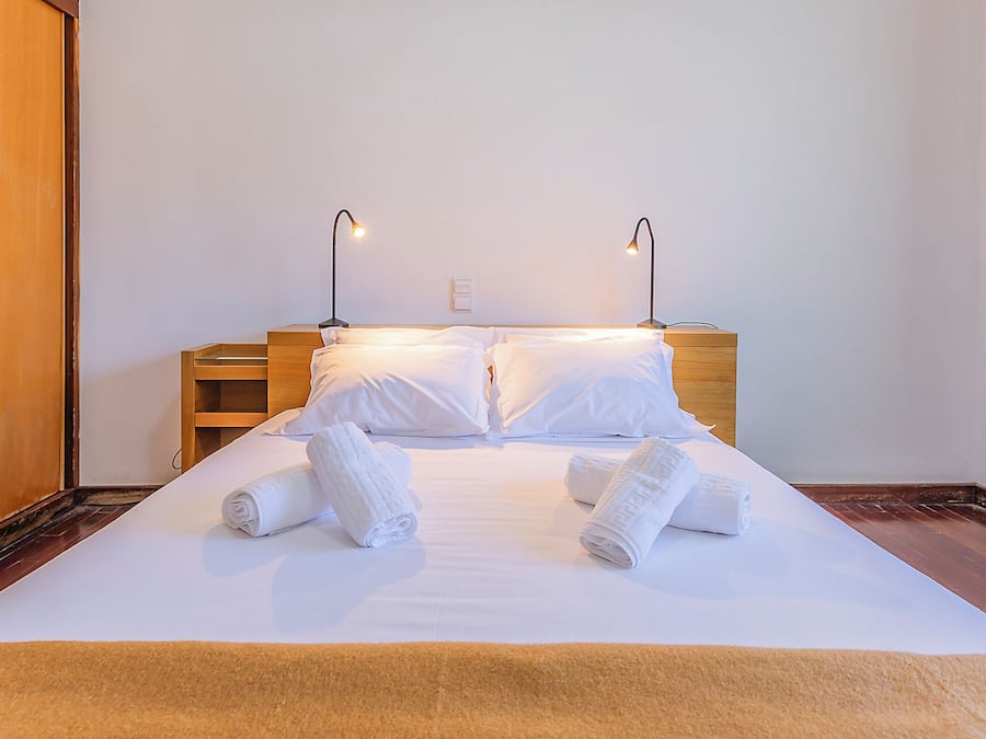 A neatly made bed with white linens and rolled towels, two bedside lamps, and a wooden headboard.