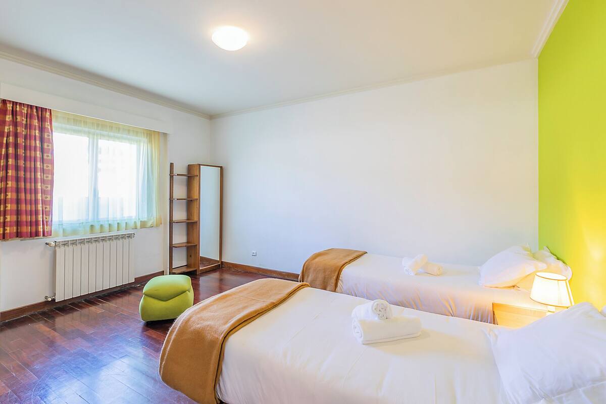 Une chambre d’hôtel avec deux lits, un radiateur, une fenêtre avec des rideaux, un pouf vert et une échelle en bois.