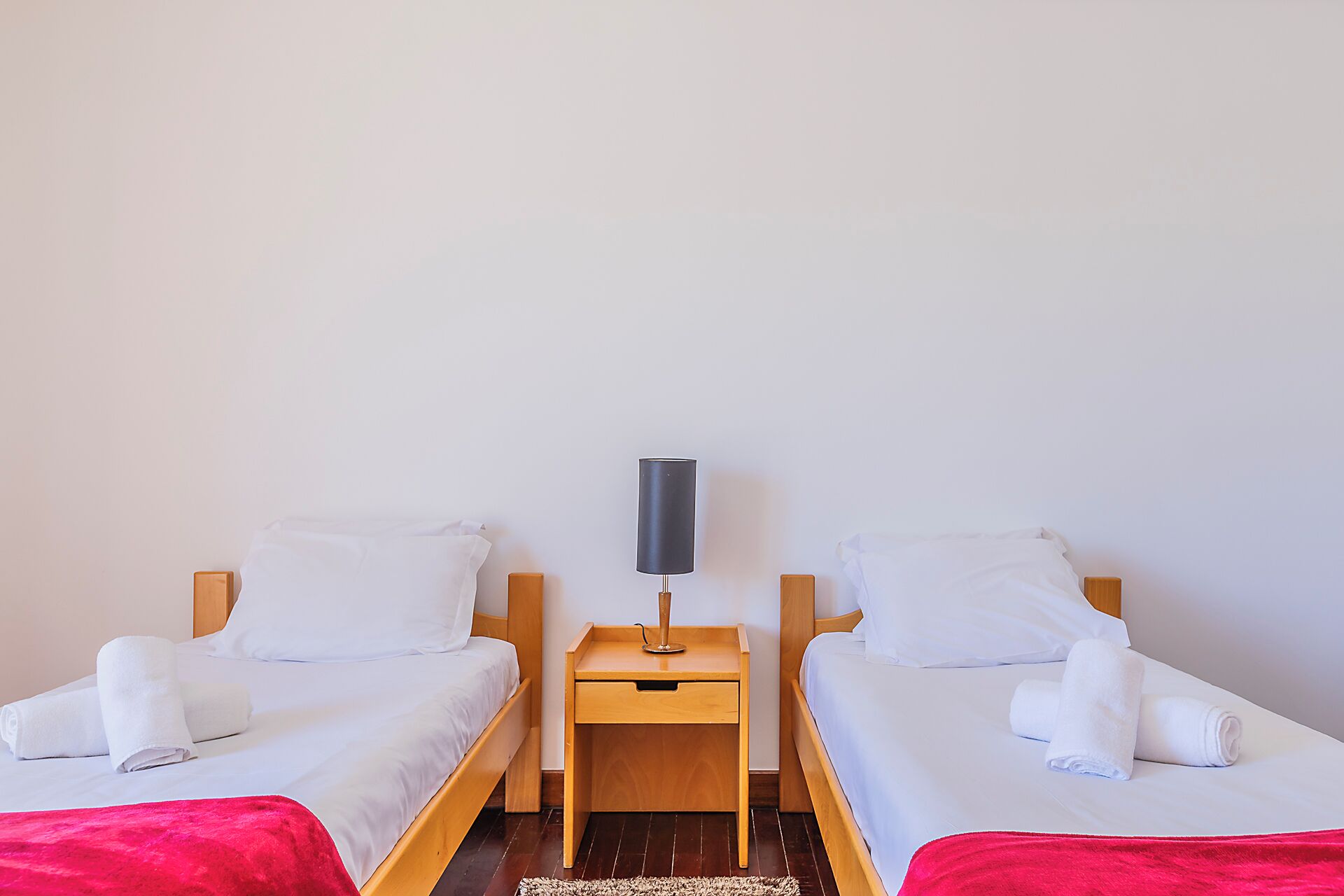 A twin room with two single beds, a bedside table with a lamp, and a red blanket on the floor.