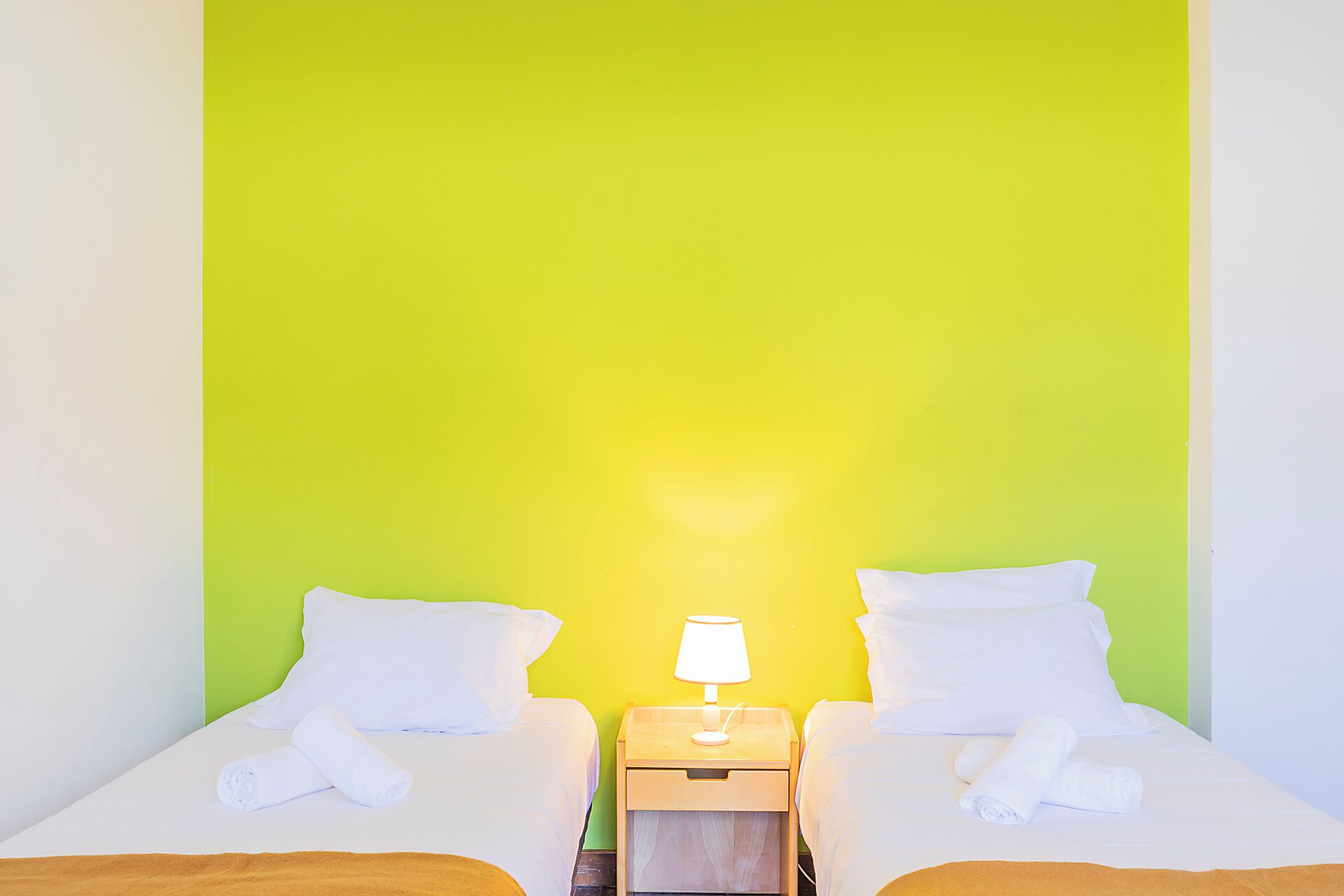 Quarto duplo com duas camas, cada uma com roupa de cama e almofadas brancas, uma mesa de cabeceira com abajur e uma parede verde de destaque.