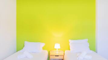 Quarto duplo com duas camas, cada uma com roupa de cama e almofadas brancas, uma mesa de cabeceira com abajur e uma parede verde de destaque.